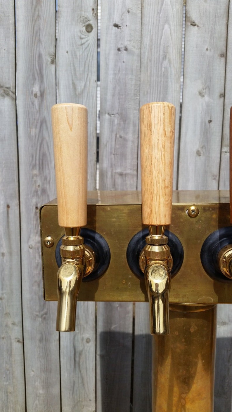 Puede incluir: Un grifo de cerveza de metal dorado con tres asas de madera. Las asas son de color marr&oacute;n claro y tienen una veta de madera natural.