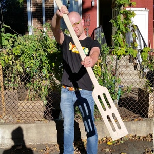 May include: A person holding a large wooden paddle with a handle and a wide, flat blade with multiple slots. The paddle is made of light-coloured wood and has a natural finish. The person is wearing blue jeans and a black t-shirt.