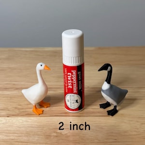 May include: A white and red lip balm tube with the text "Peppermint Twist" and two small bird figurines. One is a white goose with orange feet and beak, and the other is a gray and black goose. The lip balm is 5 cm tall.
