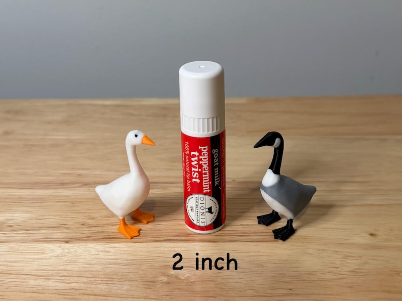 May include: A white and red lip balm tube with the text "Peppermint Twist" and two small bird figurines. One is a white goose with orange feet and beak, and the other is a gray and black goose. The lip balm is 5 cm tall.