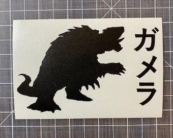 Può includere: Una silhouette nera di un mostro con i caratteri giapponesi per "Gamera" su un adesivo rettangolare bianco. Il mostro ha una schiena appuntita, artigli e una bocca aperta. L'adesivo è su una superficie a griglia grigia.