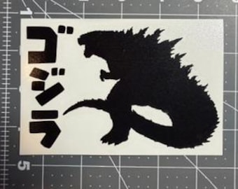 Può includere: Silhouette nera di Godzilla su un adesivo rettangolare bianco. Il mostro è in posizione eretta. I caratteri giapponesi sono impilati verticalmente a sinistra. L'adesivo è su uno sfondo a griglia grigia.