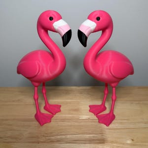 Puede incluir: Dos figuras de flamencos de color rosa brillante colocadas una al lado de la otra. Cada ave presenta un cuello largo y curvado, un pequeño ojo negro y un pico con secciones blancas, rosa claro y negras. Tienen patas finas y palmeadas.