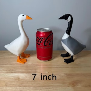 May include: Two goose ornaments, one white with orange beak and feet, the other grey, black, and white, stand on a wooden surface. A red Coca-Cola Zero Sugar can is between them. The text "7 inch" is at the bottom (approx. 18 cm).
