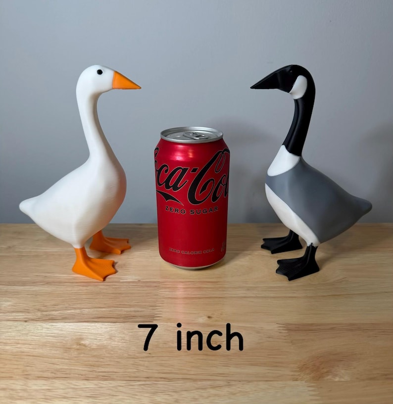 May include: Two goose ornaments, one white with orange beak and feet, the other grey, black, and white, stand on a wooden surface. A red Coca-Cola Zero Sugar can is between them. The text "7 inch" is at the bottom (approx. 18 cm).