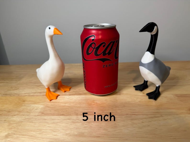 May include: Two decorative goose figurines, one white with orange beak and feet, the other gray, black, and white. A red Coca-Cola Zero can is between them. The text "5 inch" (approx. 13 cm) is at the bottom.
