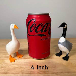 May include: Two goose figurines, one white with orange beak and feet, the other gray, black, and white, stand beside a red Coca-Cola Zero can. The can is approximately 10 cm tall. The figurines are on a wooden surface.