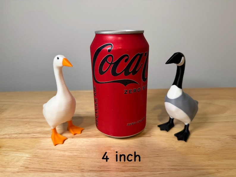 May include: Two goose figurines, one white with orange beak and feet, the other gray, black, and white, stand beside a red Coca-Cola Zero can. The can is approximately 10 cm tall. The figurines are on a wooden surface.