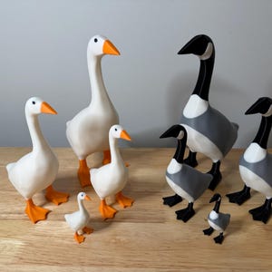 May include: A collection of goose figurines in various sizes. The geese are primarily white with orange beaks and feet. Some geese have gray and black markings. The figurines are arranged on a wooden surface against a neutral background.