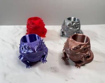 Può includere: Quattro fioriere a forma di drago barbuto stampate in 3D, nei colori rosso, argento, viola e bronzo. Ogni fioriera presenta una testa e un corpo di drago dettagliati, con un'apertura cilindrica per piante o piccoli oggetti. Le fioriere sono disposte su una superficie a motivi marmorei bianchi e grigi.