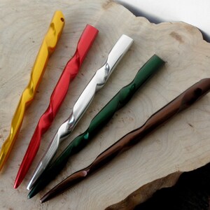 May include: Five decorative hair sticks in gold, red, silver, green, and brown. Each stick has a twisted design and a pointed end. The sticks are arranged on a light-colored wooden surface.