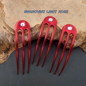 May include: Three red hair combs with a clear crystal at the top of each comb. The text "SWAROVSKI LIGHT ROSE" is printed above the combs.