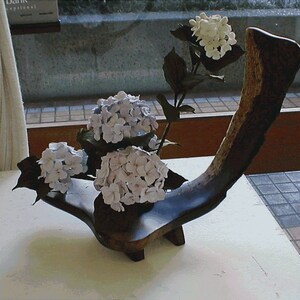 May include: A wooden vase in the shape of a curved branch holds three clusters of white hydrangeas. The vase is sitting on a white table.
