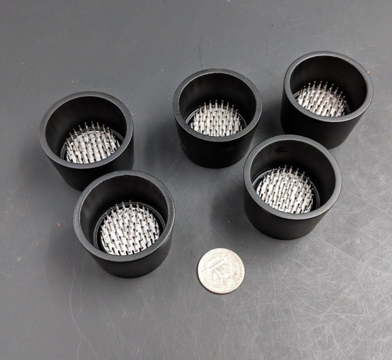May include: Five black plastic floral frog bases with silver metal pins. The bases are round and have a diameter of approximately 3 inches.