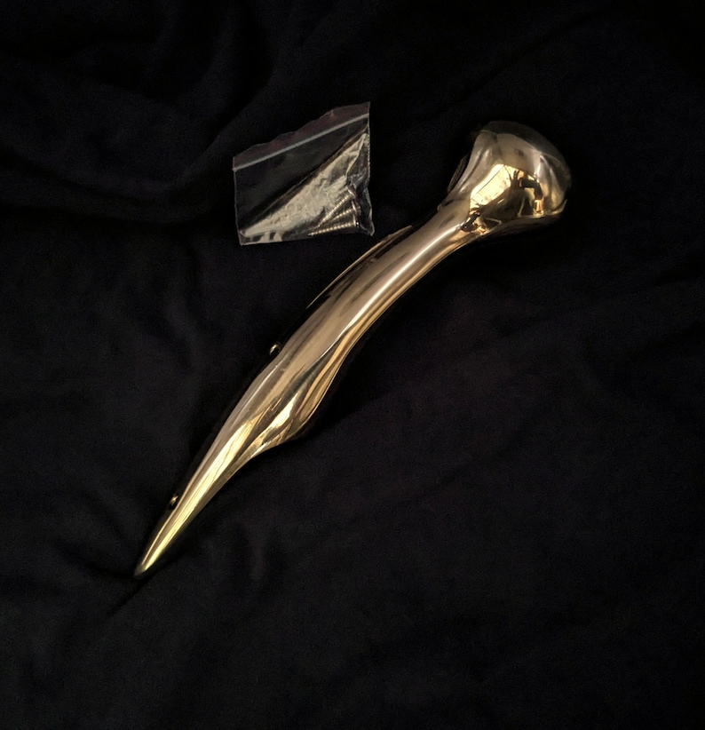 Puede incluir: Una manija de metal dorado con un extremo redondeado y una forma curva. La manija est&aacute; sobre un fondo de tela negro y viene con tornillos en una peque&ntilde;a bolsa de pl&aacute;stico.