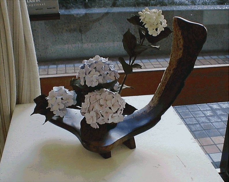 May include: A wooden vase in the shape of a curved branch holds three clusters of white hydrangeas. The vase is sitting on a white table.