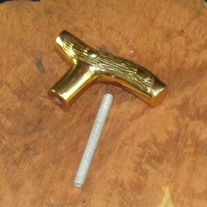 May include: A gold-colored metal cane handle with a detailed wood grain design. The handle is shaped like a tree branch and has a threaded hole for attaching a cane shaft.