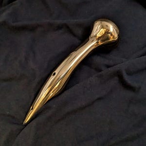 May include: A polished gold-colored metal handle with a rounded end and a pointed tip. The handle has three small holes drilled into it.