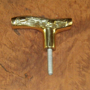 May include: A gold-colored metal T-shaped handle with a woodgrain design. The handle has a threaded shaft.