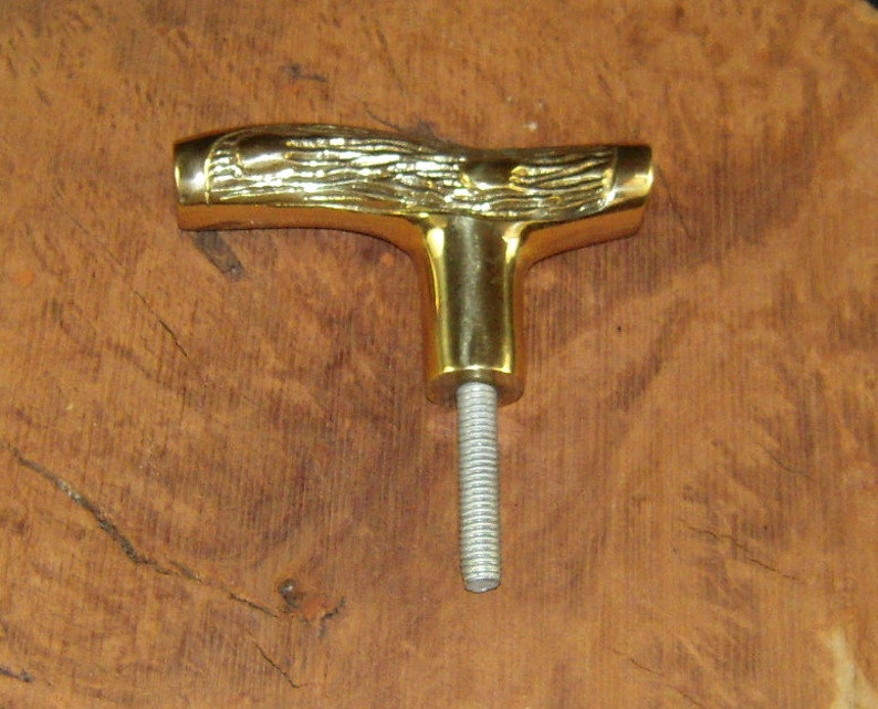 May include: A gold-colored metal T-shaped handle with a woodgrain design. The handle has a threaded shaft.