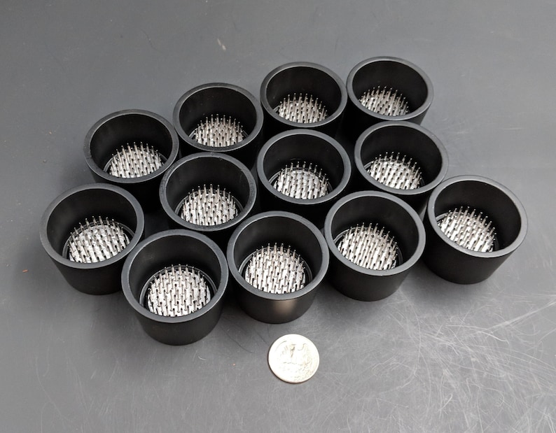 May include: Twelve black plastic cups with a circular base and a silver metal pincushion insert. The cups are arranged in a semi-circle on a gray surface.