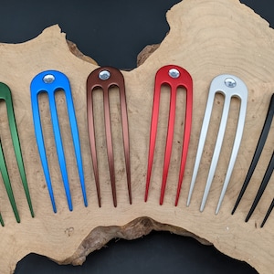 May include: Six hair combs with three tines each. The combs are made of metal and have a shiny finish. The colors of the combs are green, blue, brown, red, silver, and black. Each comb has a clear rhinestone at the top.