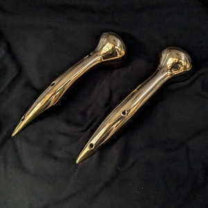 May include: Two polished gold-colored metal handles with rounded ends and a pointed tip. The handles have a smooth, shiny finish.