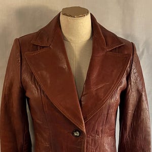 May include: A brown leather blazer featuring a wide notched collar, two front buttons, and two large patch pockets with vertical stitched detailing. The jacket has long sleeves and a textured finish, displayed on a mannequin torso.