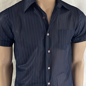 May include: Navy blue short-sleeve shirt with vertical stripes. The shirt has a button-up front with pink buttons and a chest pocket. The shirt is paired with blue jeans.