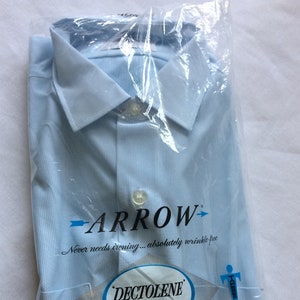 以下が含まれることがあります： 白いストライプ模様の入った薄いブルーのドレスシャツが、透明なビニール袋に入っています。シャツには「Arrow」と「Dectolene Perma-Iron」のラベルが貼られており、「アイロン不要…しわになりにくい」と「長持ち…新鮮さを保つ 100% Dacron ポリエステル」というキャッチフレーズが書かれています。
