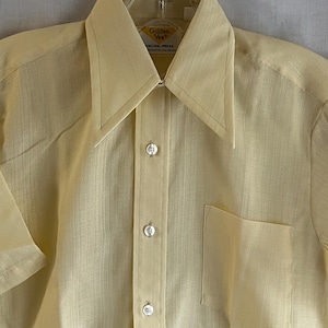 May include: A pale yellow short-sleeved button-down shirt with a pointed collar and a single chest pocket. The shirt features a vertical textured weave and white buttons. A label inside the collar reads Golden Vee Endura-Press.