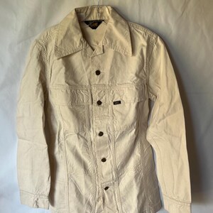 May include: Cream-coloured button-up jacket with a pointed collar and long sleeves. The jacket has two chest pockets and a small label on the right pocket. The jacket is hanging on a metal hanger.