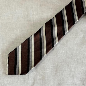 May include: A brown and black striped tie with alternating stripes of black, gray, and white. The tie is made of a smooth fabric and is angled across a neutral background.