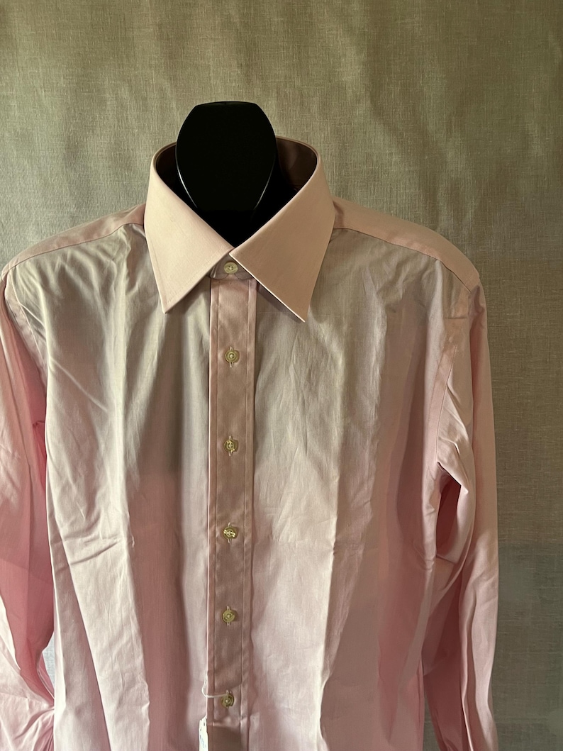 Puede incluir: Una camisa rosa claro con cuello cl&aacute;sico y mangas largas. La camisa est&aacute; abotonada en la parte delantera con botones peque&ntilde;os y redondos. La camisa se exhibe en una cabeza de maniqu&iacute; negra.