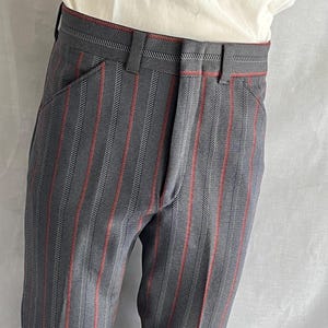 Peut inclure: Pantalon taille haute en tissu gris foncé avec un motif à rayures verticales alternant des nuances de gris et de fines lignes rouges. Le pantalon présente une ceinture structurée avec des passants et une fermeture à braguette, porté avec un haut blanc.