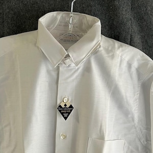 Op de afbeelding: Een wit overhemd met knoopsluiting en puntkraag, hangend aan een metalen hanger. Het overhemd heeft een zak en een label met de tekst "snap-tab". Het label toont ook de merknaam "Van Heusen".