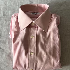 Puede incluir: Camisa rosa p&aacute;lido doblada, con cuello cl&aacute;sico y cierre abotonado. La camisa tiene un brillo sutil y est&aacute; cuidadosamente doblada, mostrando el dise&ntilde;o a medida. La etiqueta Paul Smith es visible en el cuello.