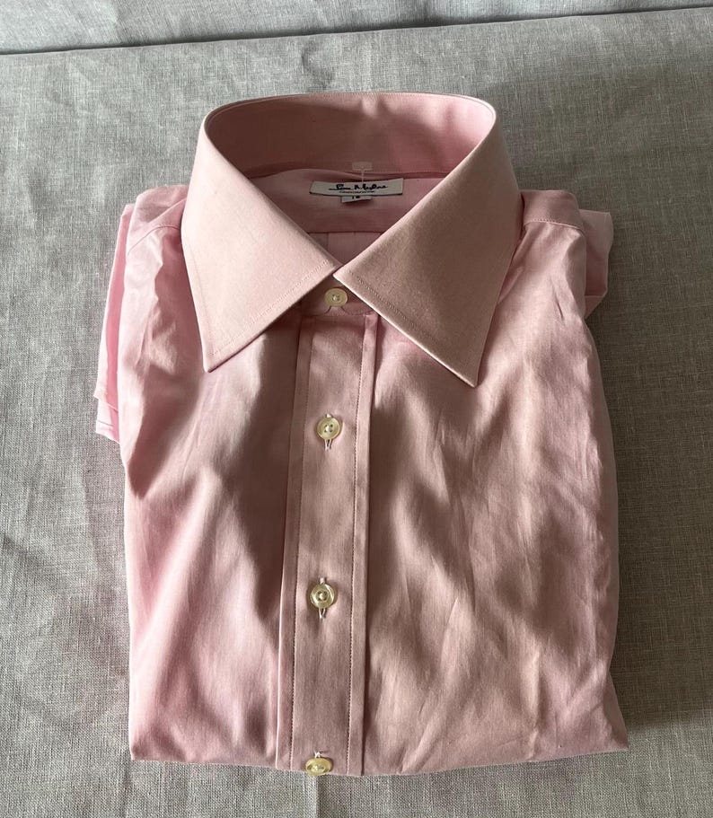 Puede incluir: Camisa rosa p&aacute;lido doblada, con cuello cl&aacute;sico y cierre abotonado. La camisa tiene un brillo sutil y est&aacute; cuidadosamente doblada, mostrando el dise&ntilde;o a medida. La etiqueta Paul Smith es visible en el cuello.