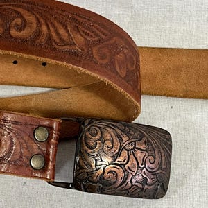 May include: A brown leather belt with a decorative copper-coloured buckle. The belt features embossed floral patterns and a natural leather finish. The buckle has an ornate design.