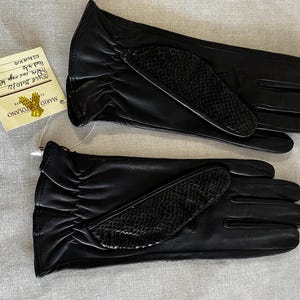Puede incluir: Un par de guantes de cuero negro con un detalle texturizado en el dorso de la mano. Los guantes tienen una etiqueta adjunta con texto escrito a mano. Los guantes son probablemente para el frío o la moda.