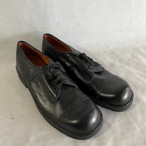 May include: A pair of black leather Oxford shoes with a rounded toe and lace-up closure. The shoes have a textured upper and a stitched sole. The shoes are displayed on a white surface.