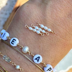 May include: Close-up of several gold bracelets and charms. One bracelet features white beads with black lettered charms spelling out "PEA". Other bracelets include pearls and gold chain links. Three small pearl and gold charms are also visible.