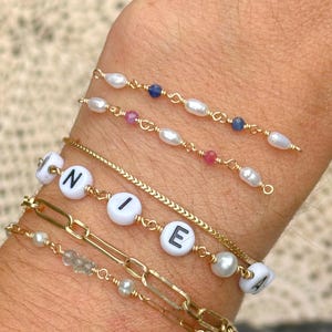 May include: Several gold bracelets with various designs are displayed. One bracelet spells out the word "NIEA" with white letter beads. Other bracelets feature pearls and small gemstones. The bracelets are on a wrist.