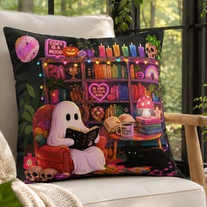 Peut inclure: Un coussin carré illustré d'un fantôme blanc assis dans un fauteuil rouge en train de lire un livre. La scène comprend une étagère avec des livres arc-en-ciel, un chat noir, une lampe champignon et le texte COZY IS A MOOD écrit en lettres.
