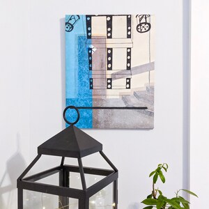 Puede incluir: Una linterna de metal negro con una carcasa de vidrio y una cadena de luces blancas en su interior. La linterna est&aacute; sentada en una mesa frente a una pared blanca con una pintura abstracta enmarcada de un edificio con un esquema de color azul y marr&oacute;n.