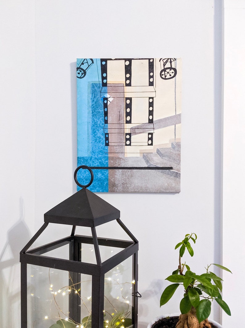 Puede incluir: Una linterna de metal negro con una carcasa de vidrio y una cadena de luces blancas en su interior. La linterna est&aacute; sentada en una mesa frente a una pared blanca con una pintura abstracta enmarcada de un edificio con un esquema de color azul y marr&oacute;n.