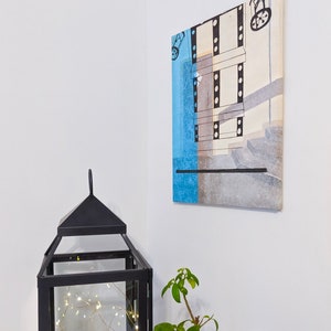 Puede incluir: Una linterna de metal negro con una caja de vidrio que contiene una cadena de luces blancas y follaje verde. Una peque&ntilde;a planta en maceta con hojas verdes est&aacute; a la derecha de la imagen. Una pintura abstracta enmarcada con colores azul, gris y blanco cuelga de la pared detr&aacute;s de la linterna.
