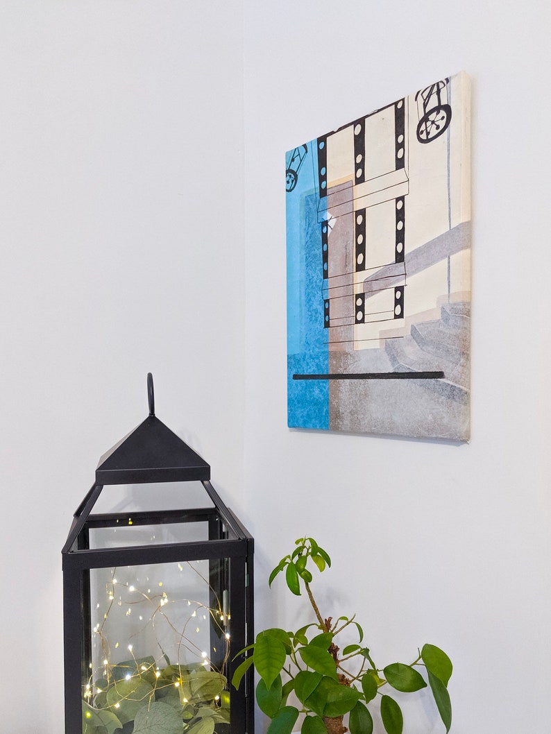 Puede incluir: Una linterna de metal negro con una caja de vidrio que contiene una cadena de luces blancas y follaje verde. Una peque&ntilde;a planta en maceta con hojas verdes est&aacute; a la derecha de la imagen. Una pintura abstracta enmarcada con colores azul, gris y blanco cuelga de la pared detr&aacute;s de la linterna.