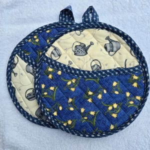 May include: Two round quilted pot holders. One features a blue floral pattern, the other a cream background with gray watering cans. Both have blue trim and hanging loops. A charming addition to any kitchen.