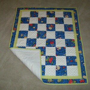 Könnte beinhalten: Eine blaue und weiße karierte Babydecke mit gelbem Rand. Die Decke besteht aus Quadraten mit verschiedenen Mustern, darunter ein Muster mit kleinen Spielzeugen.